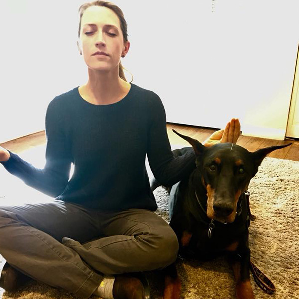 A woman sits cross-legged on the floor with her eyes closed and hands in a meditative pose. Next to her, a Doberman dog, recently examined by a veterinarian, sits calmly on the carpet with its ears perked up. The background includes a light-colored wall and a door.