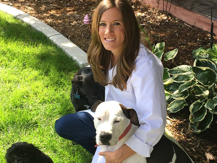 A smiling veterinarian in a white coat kneels on a green lawn, surrounded by three dogs. She holds a black and white dog in her arms, while the other two dogs, one black and one dark brown, are close by. A garden with leafy plants is visible in the background.