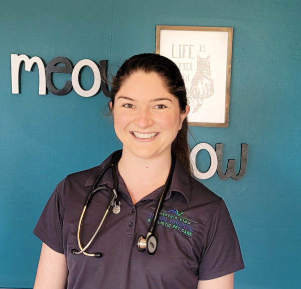 A smiling veterinarian in a dark gray polo shirt with a veterinary logo and a stethoscope around her neck stands in front of a teal wall. The wall features black-and-white cutout letters spelling "meow" and a framed sign that reads "Life is better with cats.