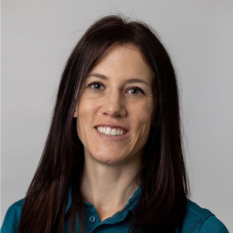 A woman with long, dark brown hair is smiling against a plain light gray background. She is wearing a teal collared shirt, giving off the professional vibe of a dedicated veterinarian.