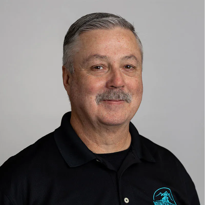A middle-aged man with short gray hair and a mustache is smiling. He is wearing a black polo shirt with a blue embroidered logo, emblematic of his profession as a dedicated veterinarian. The background is plain and light-colored.