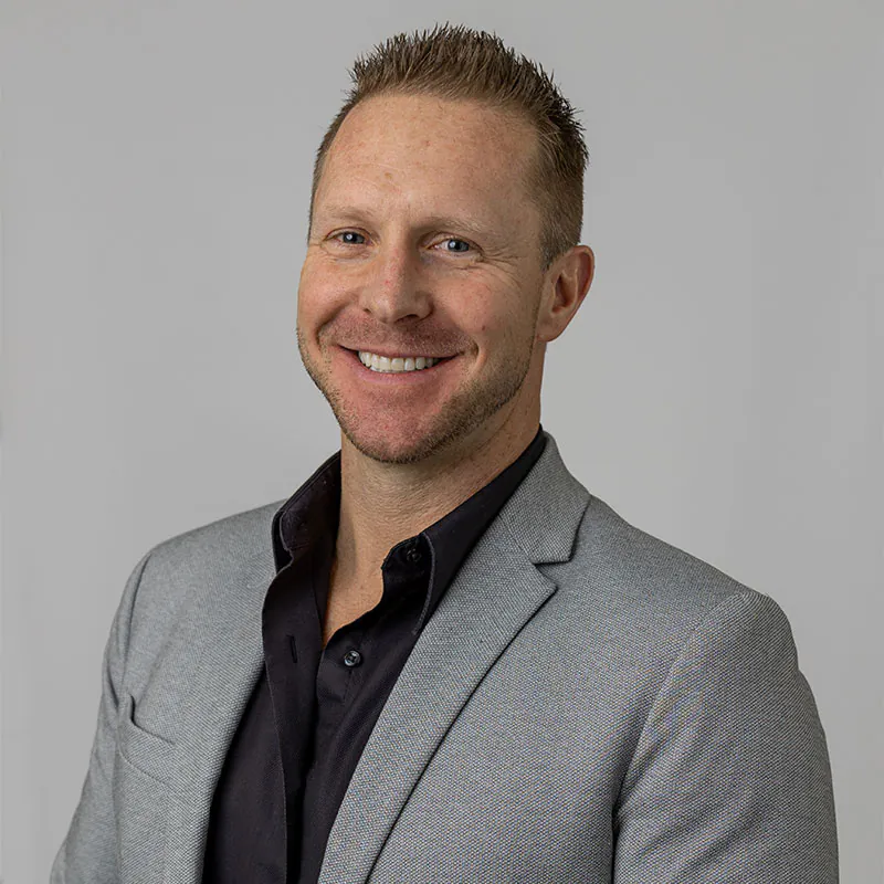 A veterinarian with short, light-colored hair smiles while posing for a professional headshot. He is wearing a light gray blazer over a black collared shirt. The background is a plain, light gray.