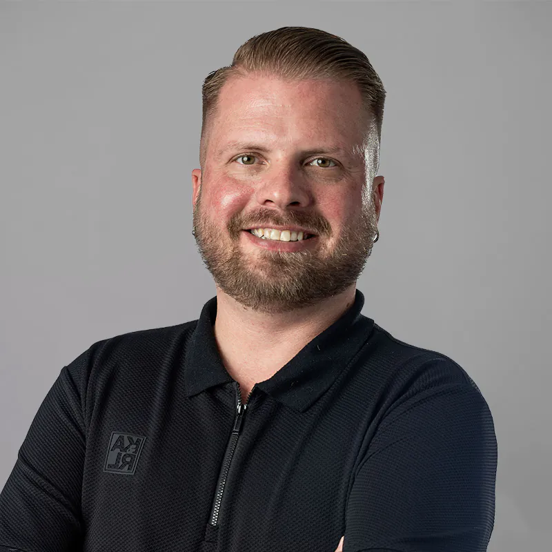 A man with short, neatly styled hair, a beard, and a smiling expression stands against a neutral gray background. He is wearing a dark polo shirt with a small logo on the upper left side that subtly hints at his profession as a veterinarian. His arms are crossed over his chest.