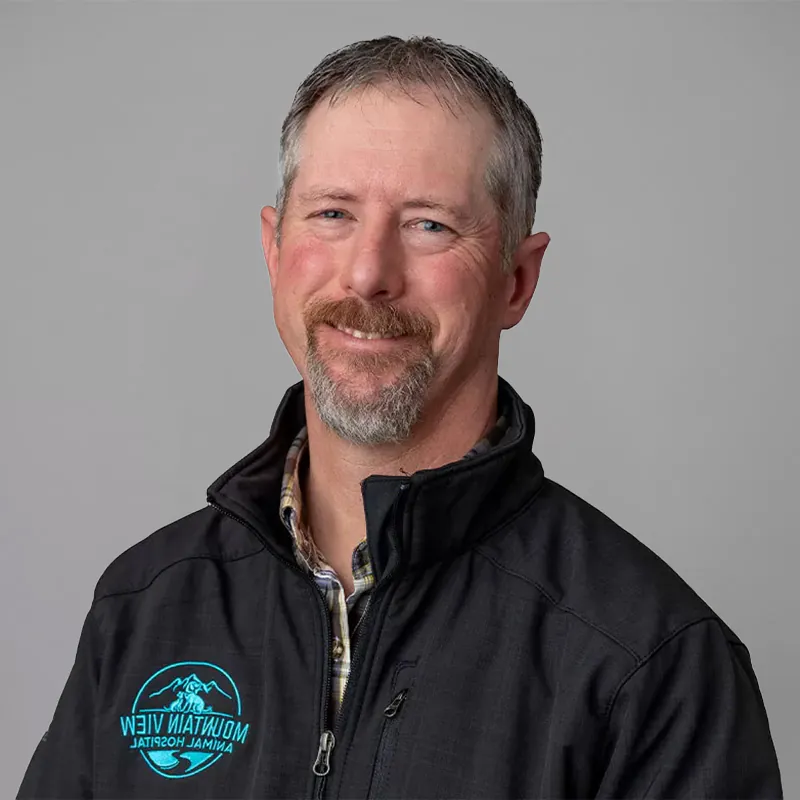 A man with short gray hair and a beard smiling at the camera. He is wearing a black jacket with a "Mountain View Animal Hospital" logo and a checkered shirt underneath, reflecting his role as a dedicated veterinarian. The background is plain gray.