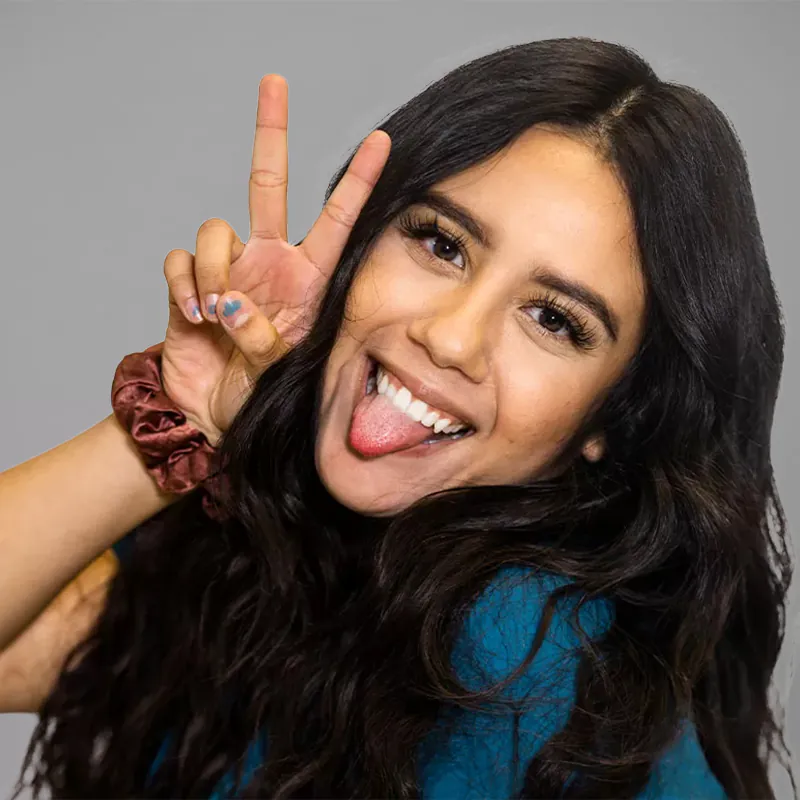 A person with long dark hair smiles broadly while sticking out their tongue and flashing a peace sign with their left hand. They are wearing a blue top and a pink scrunchie on their wrist, posed against a plain gray background, exuding the playful spirit of your favorite vet.