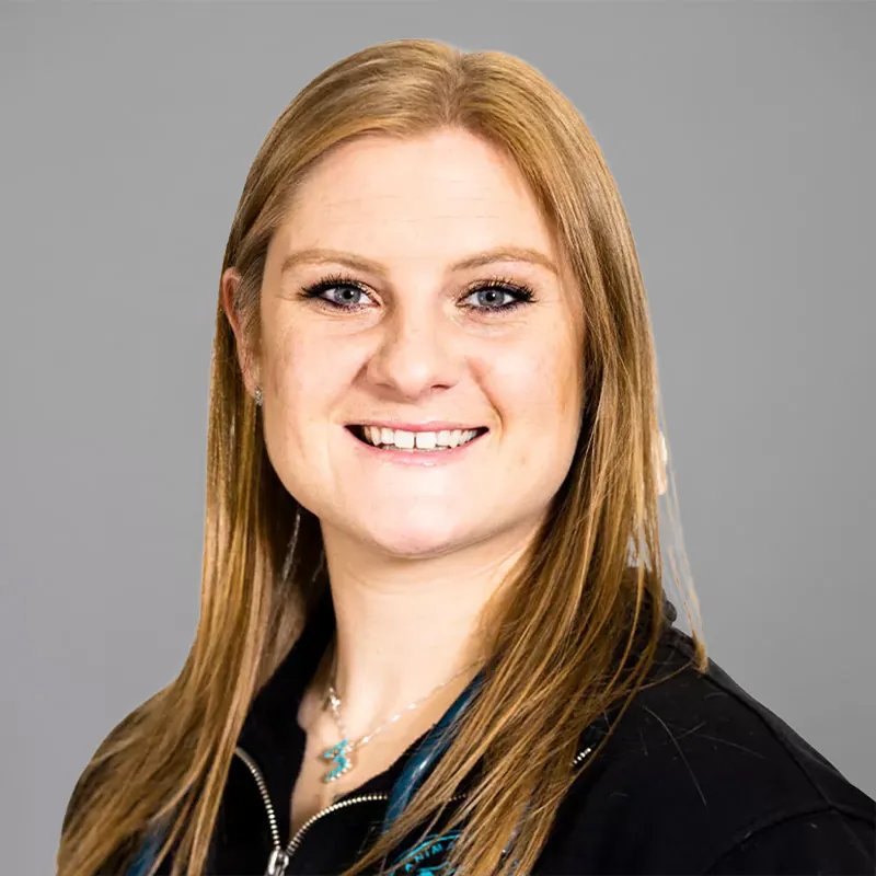 Portrait of a smiling person with long, straight blonde hair and light skin, wearing a black top and a necklace. The background is gray, giving an air of professionalism that complements the veterinarian's warm demeanor.