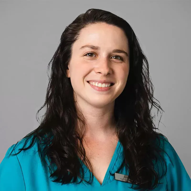 A woman with long dark hair wearing a teal scrub top and a name tag, standing against a plain gray background, smiling at the camera. It's clear she’s a veterinarian dedicated to caring for animals.