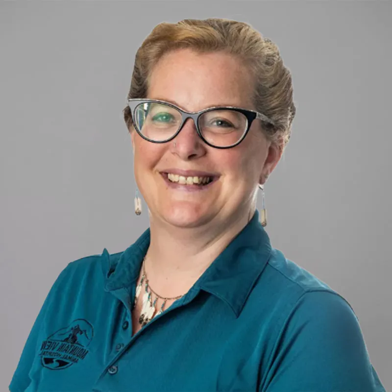 A smiling person with short, light brown hair is wearing black-framed glasses, light-colored earrings, and a teal polo shirt with a veterinarian logo on it. The background is a plain, light gray color.
