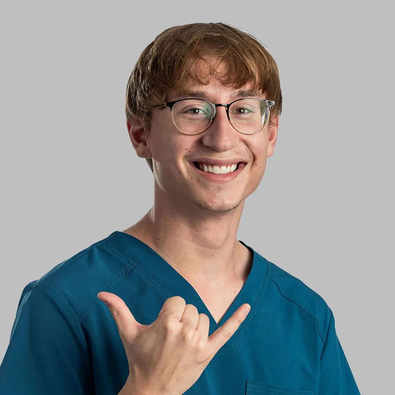 A young person with short light brown hair and glasses is smiling and holding up a shaka sign with their right hand. They are wearing blue medical scrubs, embodying the perfect image of a dedicated veterinarian, standing against a plain gray background.