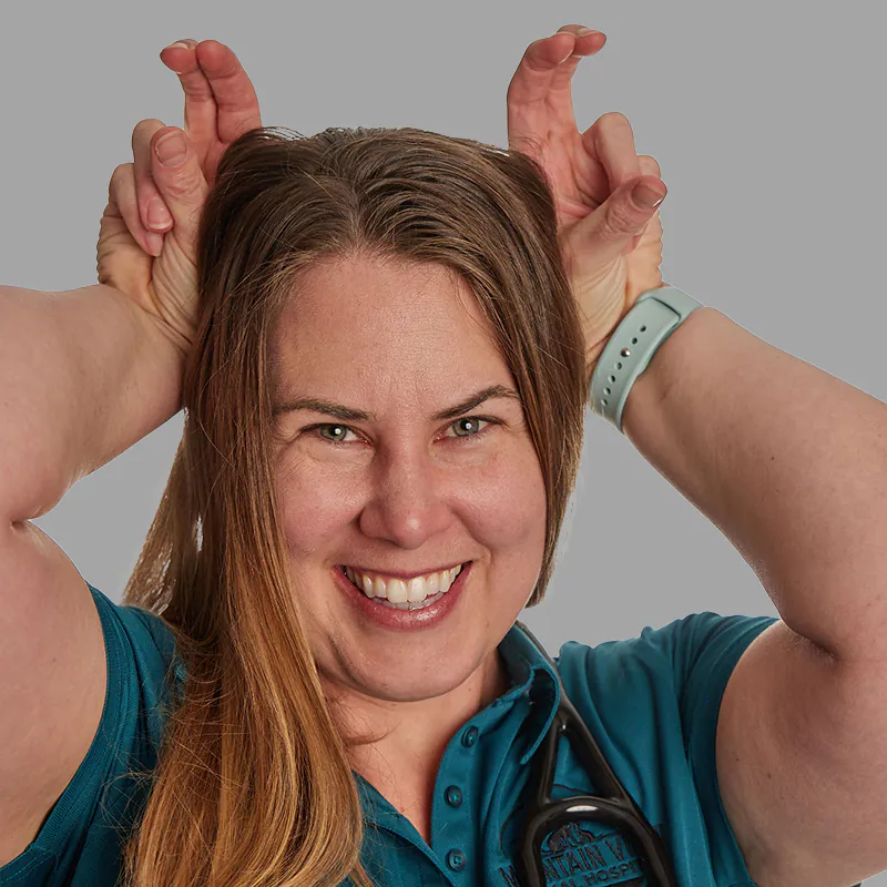 A woman with long hair, smiling broadly, holds her hands up to her head with fingers pointed upwards, mimicking animal ears or horns. She is wearing a teal shirt and a light blue watch, with a stethoscope around her neck, embodying the playful spirit of a dedicated veterinarian. The background is a plain gray.