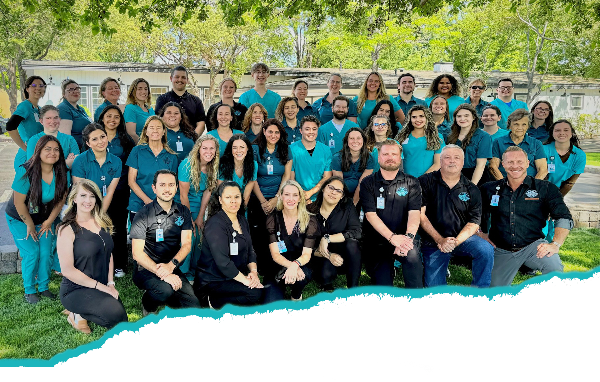 A large group of people, consisting of men and women, is posing for a photo outdoors on a grassy area. Some are wearing blue medical scrubs, indicating their roles as veterinarians, while others are dressed in black. Trees and a building are visible in the background. Everyone is smiling.