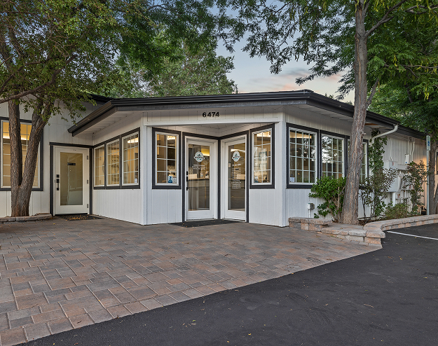 A white building with large windows, labeled with the number 6474, surrounded by trees. The entrance has a glass door and paved walkway, with a blacktop driveway in front.