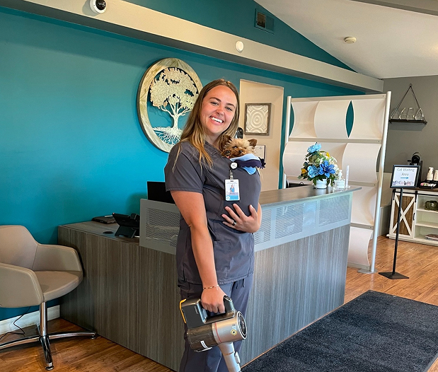 A smiling woman in scrubs stands in a modern office holding a small dog in a pouch on her chest and a handheld vacuum. A desk with a computer and blue wall décor are in the background.
