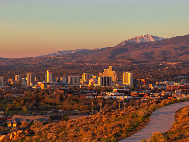 A city skyline at sunset with tall buildings, a river, and tree-lined streets, backed by rolling hills and a snow-capped mountain in the distance under a clear sky.