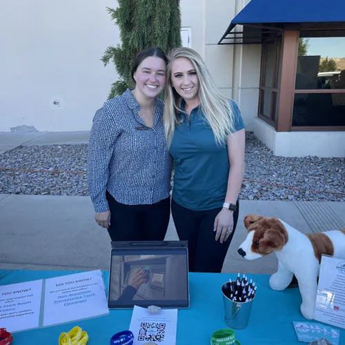 Two women stand smiling behind an outdoor informational table with flyers, pens, and a plush dog. A tablet and a QR code are displayed on the table. There are trees, rocks, and a building in the background.