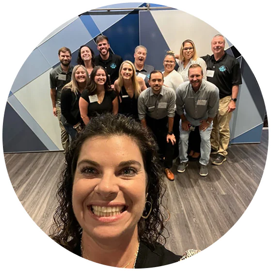 A woman in the foreground takes a smiling group selfie with eleven colleagues, all smiling, posed in front of a geometric blue and gray wall. Everyone is wearing name tags and business casual attire.