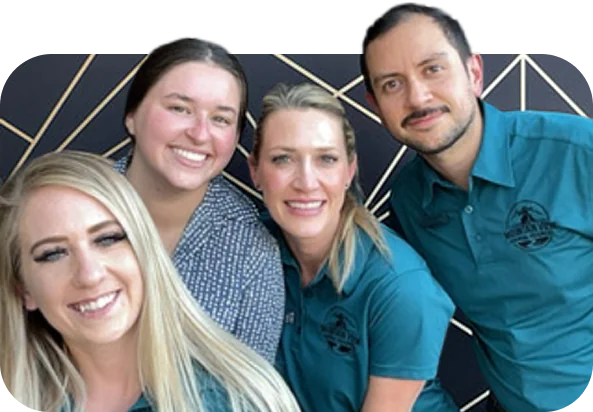 Four adults smiling and posing for a photo, three wearing matching teal shirts with a logo and one in a patterned shirt, standing in front of a geometric black and gold wall.