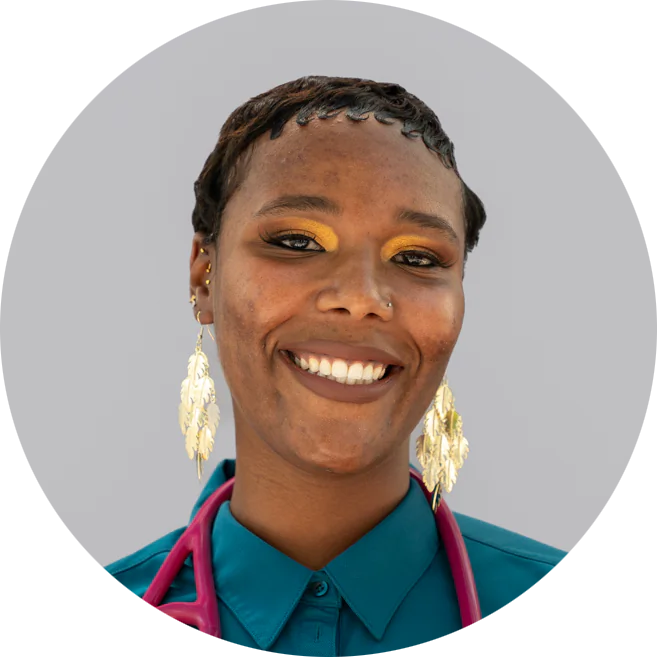 A smiling person with short hair and golden earrings, wearing a teal shirt and a pink stethoscope, stands against a plain gray background.