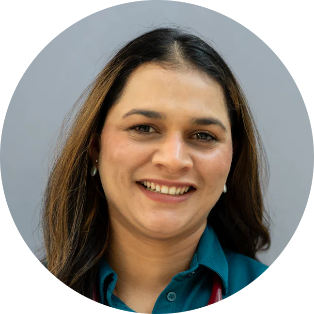 A woman with long brown hair and a teal shirt smiles at the camera against a plain gray background. She is wearing pearl earrings and a stethoscope around her neck.
