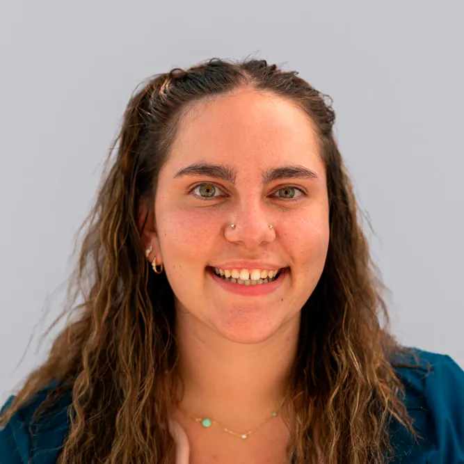 A young woman with wavy brown hair, smiling, wearing a teal top, small hoop earrings, nose piercings, and a delicate necklace, posing against a plain light gray background.