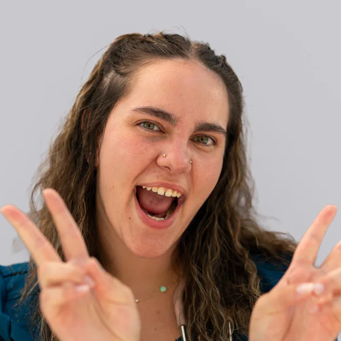 A young woman with wavy brown hair smiles widely and makes peace signs with both hands. She is wearing a blue top, has a nose piercing, and is standing in front of a plain light gray background.