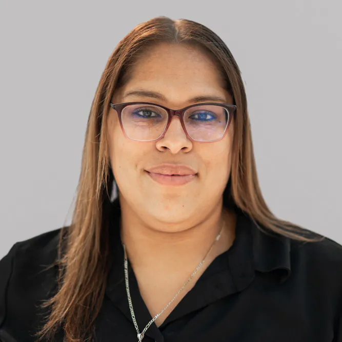 A person with straight light brown hair and glasses, wearing a black shirt and a silver necklace, smiles softly against a plain light gray background.