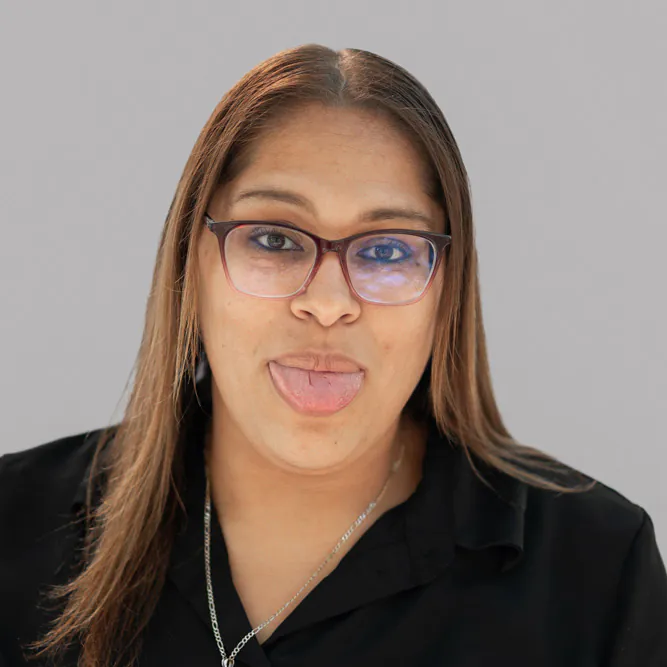 A woman with straight brown hair and glasses sticks out her tongue playfully. She is wearing a black top and has a neutral gray background.