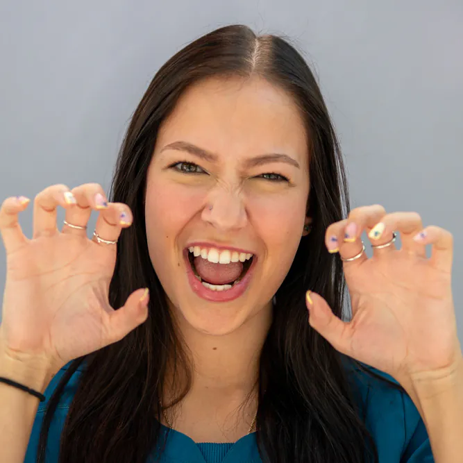 A young woman with long dark hair playfully bares her teeth and raises her hands like claws, as if pretending to roar. She is wearing rings and a blue top, smiling enthusiastically against a plain background.