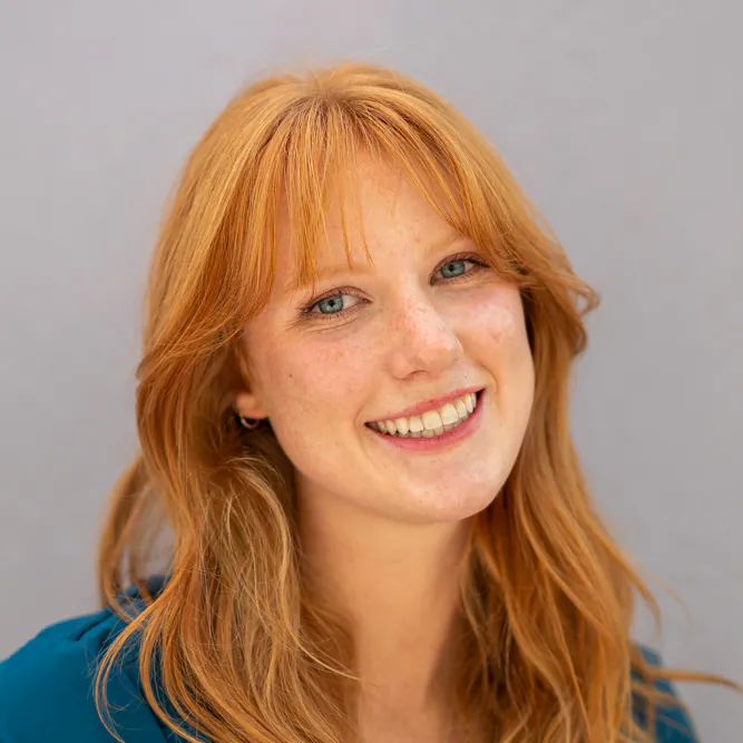 A young woman with long red hair and fair skin smiles at the camera. She is wearing a blue top and standing in front of a plain, light-colored background.