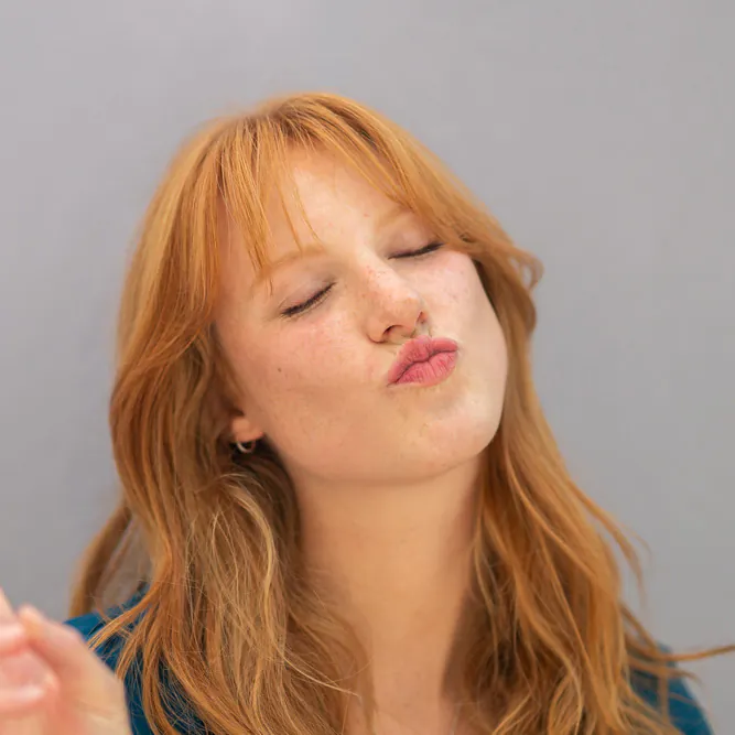 A woman with long red hair is facing the camera with her eyes closed and lips puckered, as if blowing a kiss. The background is plain and light gray.