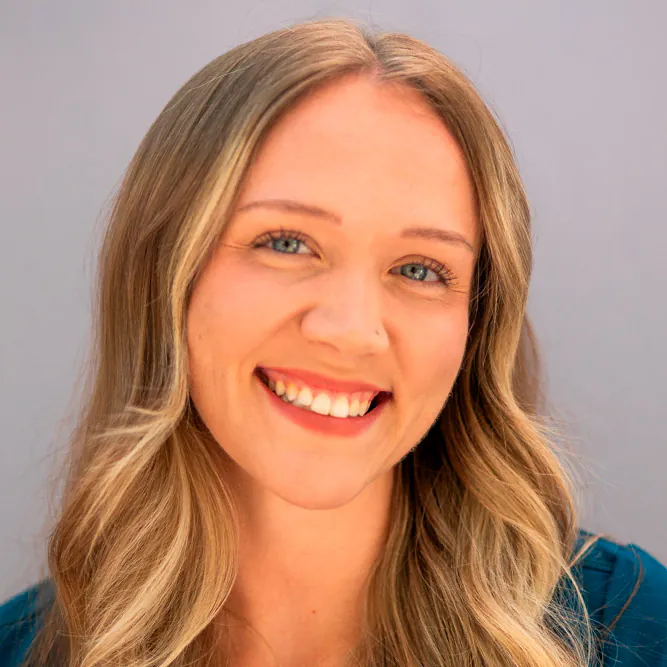 A woman with long, wavy blonde hair smiles at the camera against a plain, light-colored background. She is wearing a teal top.