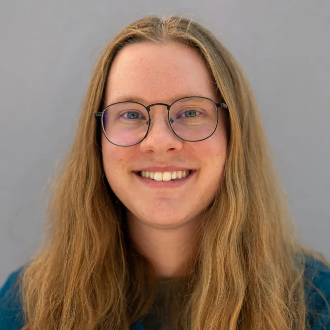 A person with long, light brown hair and glasses smiles at the camera. They are wearing a blue top and standing against a plain, light gray background.
