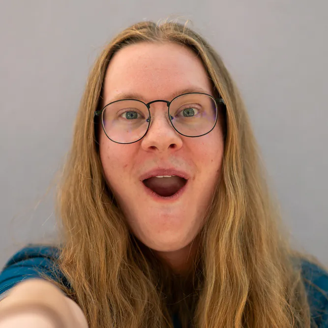 A person with long blonde hair, glasses, and a blue shirt smiles with an open mouth, looking directly at the camera against a plain light gray background.