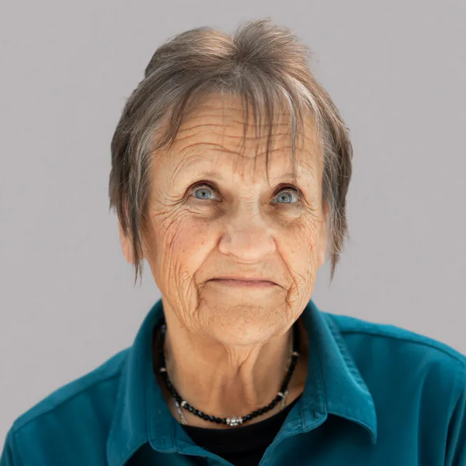 An elderly woman with short gray hair and blue eyes, wearing a teal shirt and a black beaded necklace, looks slightly upward with a gentle expression. The background is plain and light gray.