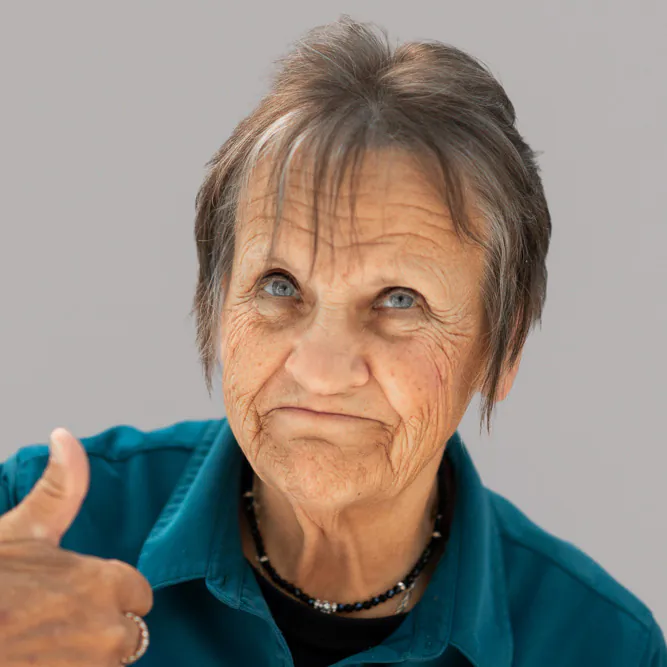 An older woman with short gray hair wearing a teal shirt gives a thumbs up. She has a neutral expression, visible wrinkles, and is wearing a beaded necklace. The background is plain and light gray.