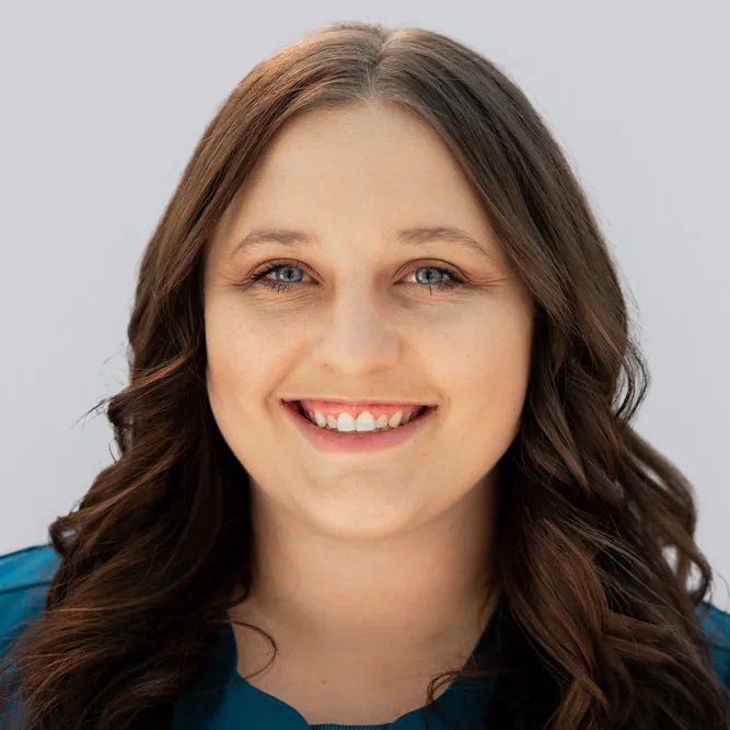A woman with long, wavy brown hair smiles at the camera. She has blue eyes and is wearing a teal top. The background is plain and light-colored.