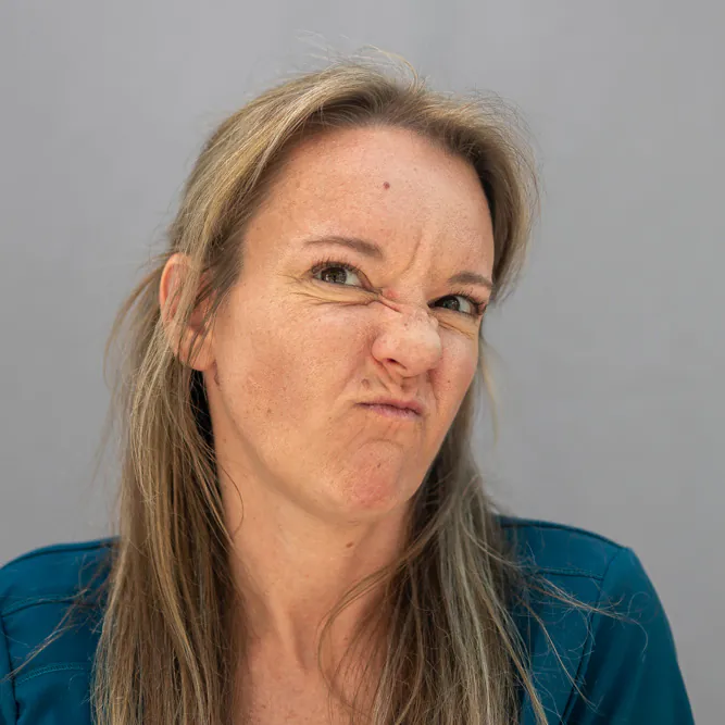 A woman with long blonde hair wearing a teal top scrunches her face in a playful or exaggerated expression, with her nose wrinkled and lips pursed, against a plain gray background.