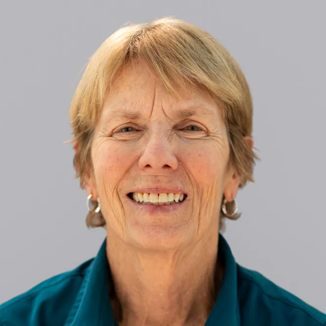 An older woman with short light brown hair and hoop earrings, smiling at the camera. She is wearing a teal collared shirt and is set against a plain light gray background.