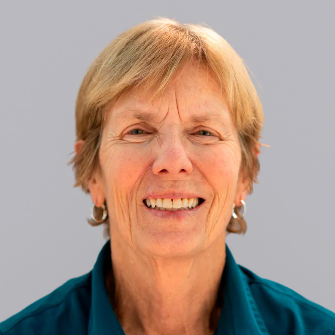 An older woman with short, light brown hair, smiling and wearing a teal shirt and silver hoop earrings, against a plain light gray background.