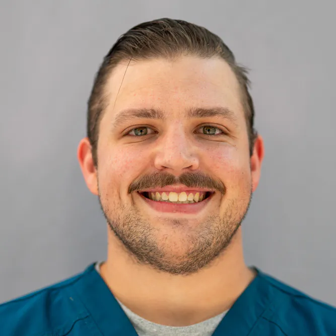 Smiling man with short brown hair and a mustache, wearing a teal scrub top and gray undershirt, standing in front of a plain light gray background.