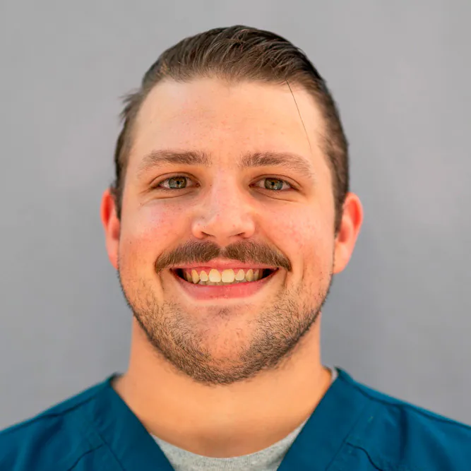 A smiling man with short brown hair and a mustache, wearing a teal scrub top, stands in front of a plain light gray background.