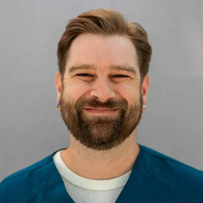 A bearded man with light brown hair and earrings is smiling. He is wearing a teal scrub top over a white shirt, standing in front of a plain, light gray background.