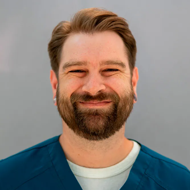 A bearded man with light brown hair and earrings smiles warmly at the camera. He is wearing teal medical scrubs over a light-colored shirt, standing against a plain gray background.