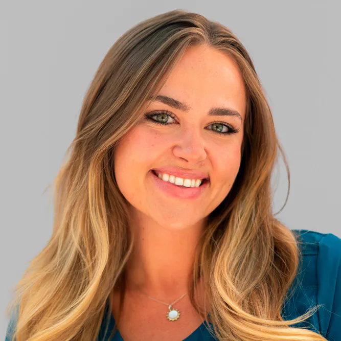 A woman with long, blonde hair and green eyes smiles at the camera. She is wearing a teal top and a pendant necklace. The background is plain light gray.
