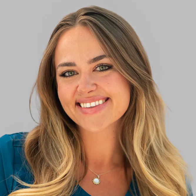 A woman with long, blonde hair and blue eyes smiles at the camera. She is wearing a teal top and a necklace with a round pendant. The background is a plain light gray.