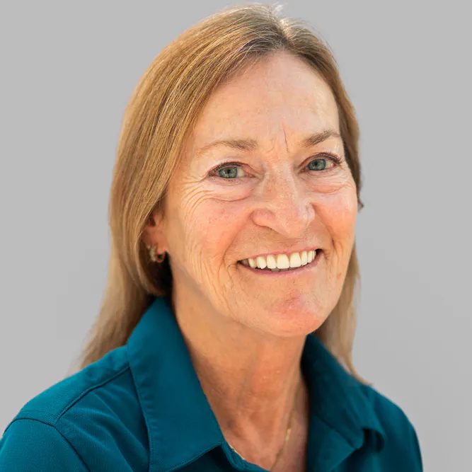 Smiling middle-aged woman with light brown hair wearing a teal collared shirt, posing against a plain light gray background.
