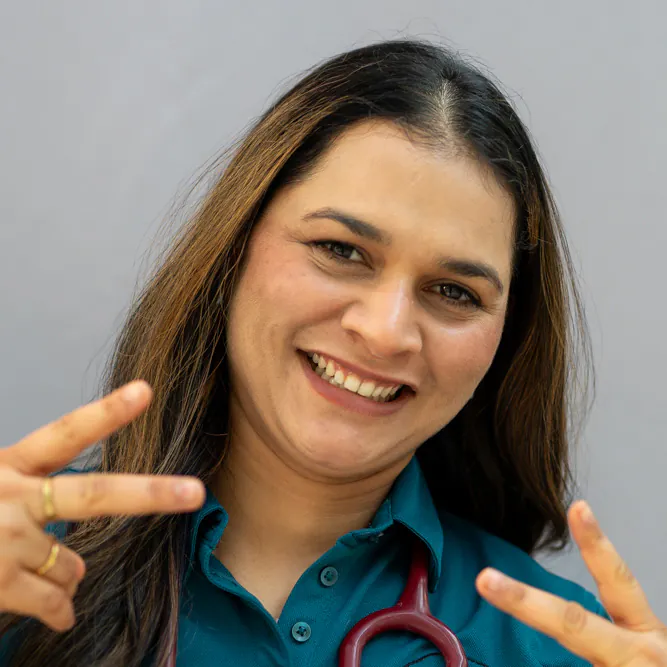 A smiling woman with long brown hair, wearing a teal shirt and a stethoscope, holds up both hands making peace signs against a plain light background.