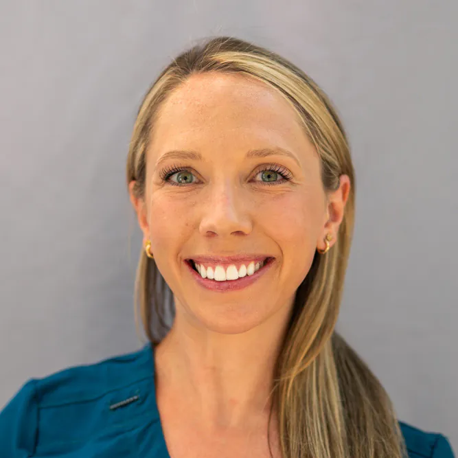 A woman with long blonde hair, wearing a teal top and small gold hoop earrings, smiles at the camera in front of a plain light gray background.