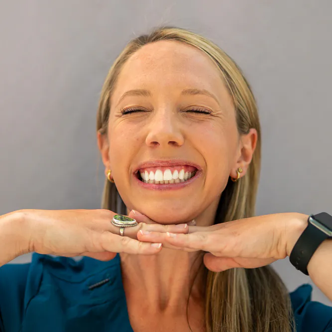 A woman with long blonde hair smiles brightly with her eyes closed, resting her chin on her hands. She wears a blue top, gold earrings, a ring, and a smartwatch, and stands against a plain gray background.
