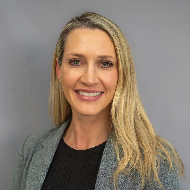 A woman with long blonde hair smiles at the camera. She is wearing a grey blazer over a black top and stands in front of a plain light grey background.
